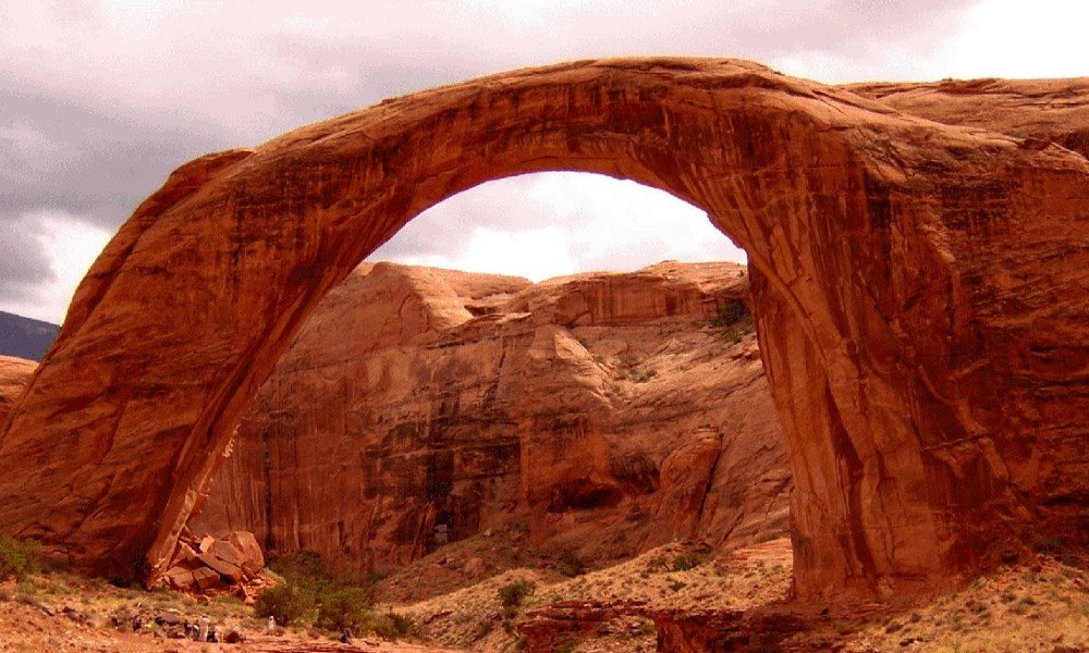 A large, natural stone arch rises above desert terrain with rugged, reddish rock formations under a cloudy sky—the perfect backdrop for those seeking lodging in Kanab UT while exploring the area’s breathtaking landscapes.