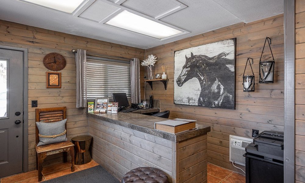 A rustic office with wood-paneled walls, a gray front desk, a computer monitor, horse artwork, brochures highlighting lodging in Kanab UT, a chair with a cushion, and a window with gray curtains. Lanterns and a clock decorate the space.