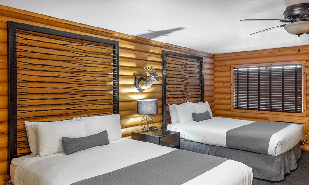 A cozy log cabin-style bedroom, perfect for lodging in Kanab UT, features two beds with gray and white bedding, a nightstand with a lamp, a deer head wall decoration, warm wood walls, and a ceiling fan with a light.