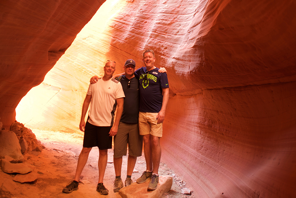 Three men stand together smiling inside a sunlit, narrow red rock canyon with smooth, curved walls. The men wear casual shorts and t-shirts, posing on sandy ground with arms around each other—perhaps celebrating their latest adventure as members of the Canyons Lodge rewards program.