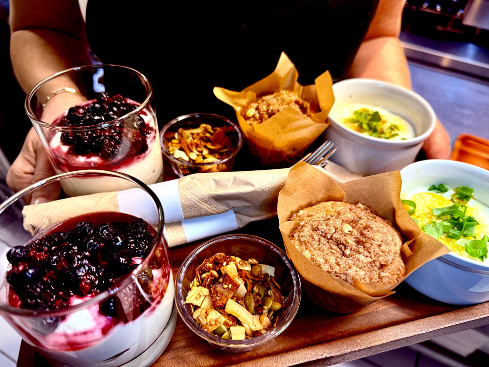 A person holds a tray with parfaits topped with berries, muffins in paper wrappers, small bowls of green-topped soup, and granola in cups, along with napkins and utensils—an inviting breakfast spread often enjoyed by guests lodging in Kanab UT.