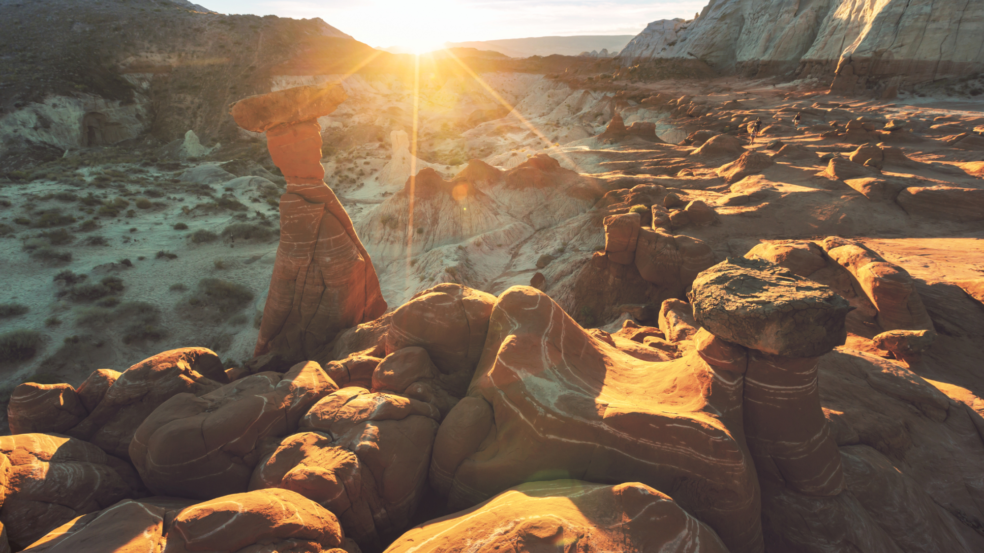 Sunlight shines over a rocky desert landscape with hoodoos and sparse vegetation, highlighting the textures and shapes of the formations.